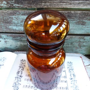 May include: A vintage amber glass jar with a rounded lid. The jar is empty and has a smooth, shiny surface. The jar is sitting on a sheet of music.