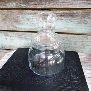 May include: A clear glass jar with a round base and a spherical lid. The jar sits on a black, textured book with an embossed design. The background features weathered wooden planks.