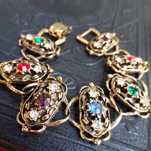 May include: Vintage gold-toned bracelet with oval links, each adorned with colorful gemstones. The bracelet features red, green, blue, and purple stones, and clear crystals. The clasp is also gold-toned.
