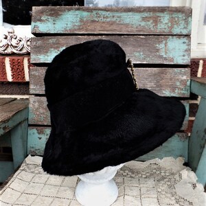 May include: A black wide-brimmed hat with a black velvet band and a gold-toned decorative pin. The hat is sitting on a white pedestal.