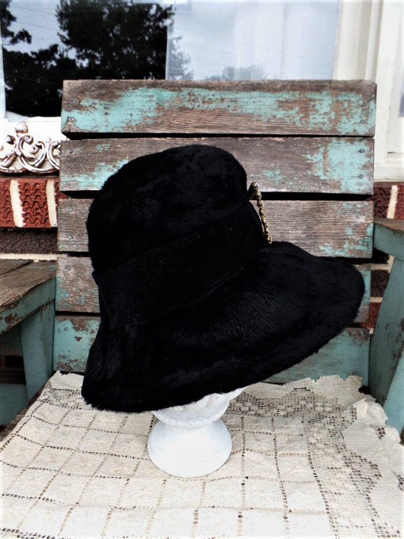 May include: A black wide-brimmed hat with a black velvet band and a gold-toned decorative pin. The hat is sitting on a white pedestal.