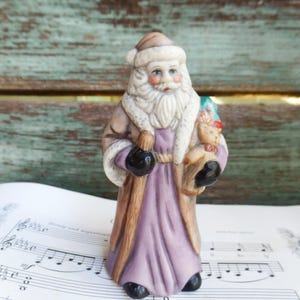 May include: A ceramic figurine of Santa Claus holding a bag of toys. He is wearing a purple robe, a white beard, and a brown hat. The figurine is standing on a sheet of music.