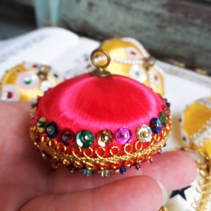 May include: A bright pink satin Christmas ornament with a gold trim and colorful sequins. The ornament has a small loop at the top for hanging.