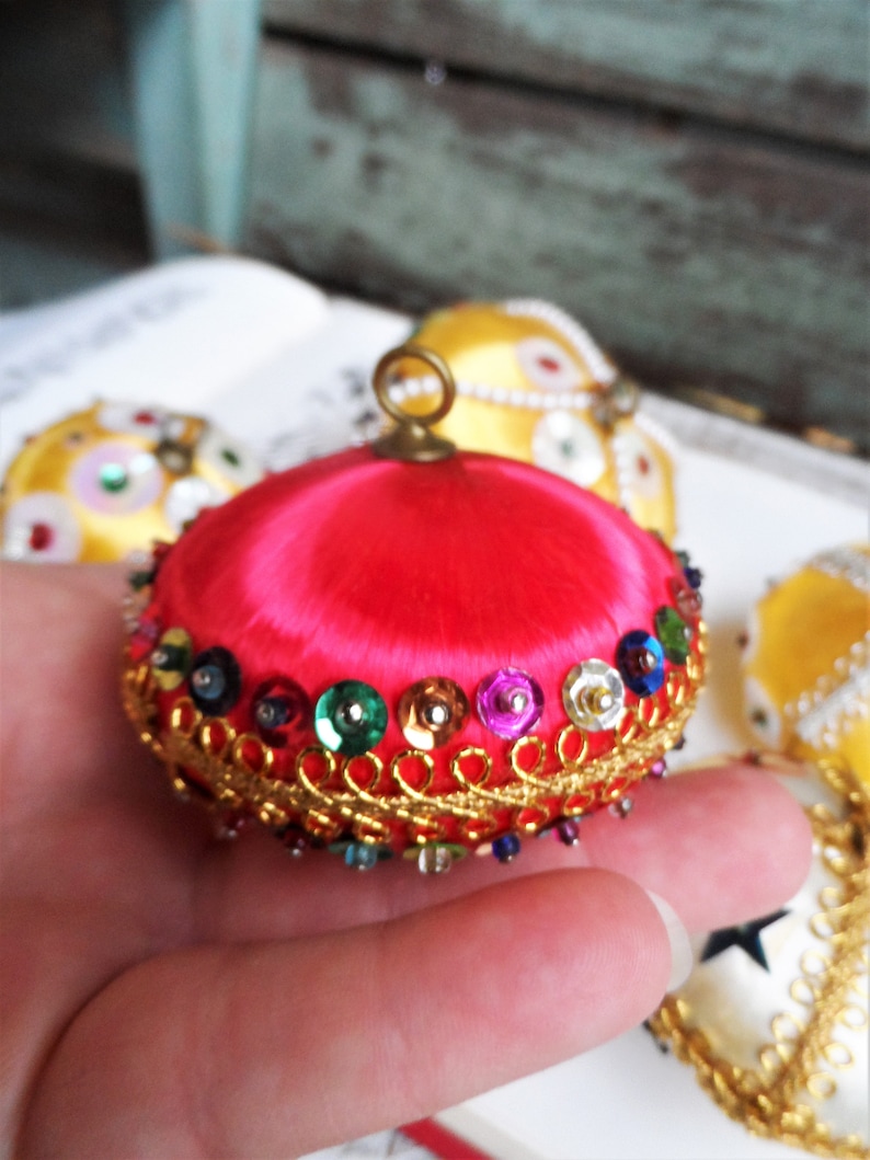 May include: A bright pink satin Christmas ornament with a gold trim and colorful sequins. The ornament has a small loop at the top for hanging.