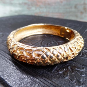 May include: A gold-colored bangle bracelet with an ornate, textured surface. The bracelet features a repeating pattern of interlocking shapes and a hinged clasp. The jewelry is displayed on a dark, patterned surface.