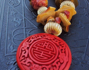 May include: A red, round pendant with a gold-colored metal bail. The pendant features a carved design of a Chinese character in a circle surrounded by floral motifs.