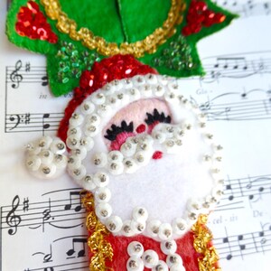 May include: A felt Santa Claus ornament featuring a white beard and hat trim embellished with clear sequins. The design includes a green holly leaf topper with gold and red sequins, and a red base with gold metallic trim and white sequin lettering.