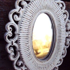 May include: White oval mirror with a decorative, ornate frame. The frame has a distressed, white paint finish and intricate scrollwork.