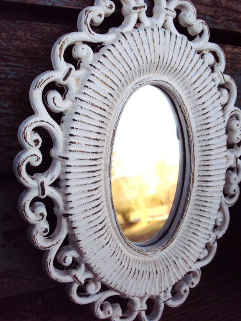 May include: White oval mirror with a decorative, ornate frame. The frame has a distressed, white paint finish and intricate scrollwork.