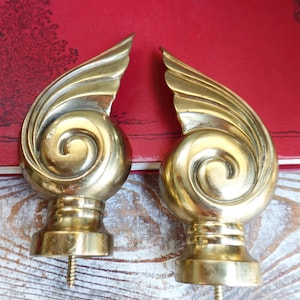 May include: Two brass-colored curtain finials with a spiral design and wing-like accents. Each finial has a round base and a screw for attachment. The background is a red book and a white-painted wooden surface.