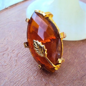 May include: A gold ring with a large, faceted, amber-colored stone. The stone is set in a gold setting with a small gold leaf design.