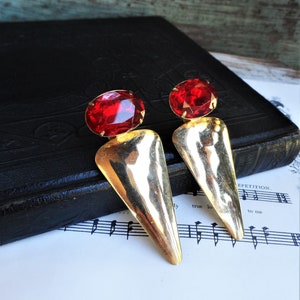 May include: A pair of gold tone earrings with red oval shaped stones. The earrings have a hammered metal design and a pointed shape.