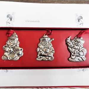 Può includere: Set di tre decorazioni natalizie argentate a forma di Babbo Natale con nastri rossi per appenderle. Gli ornamenti sono esposti in una scatola rossa con la scritta "Christmastide" stampata all'interno del coperchio. Ogni ornamento ha un design unico.