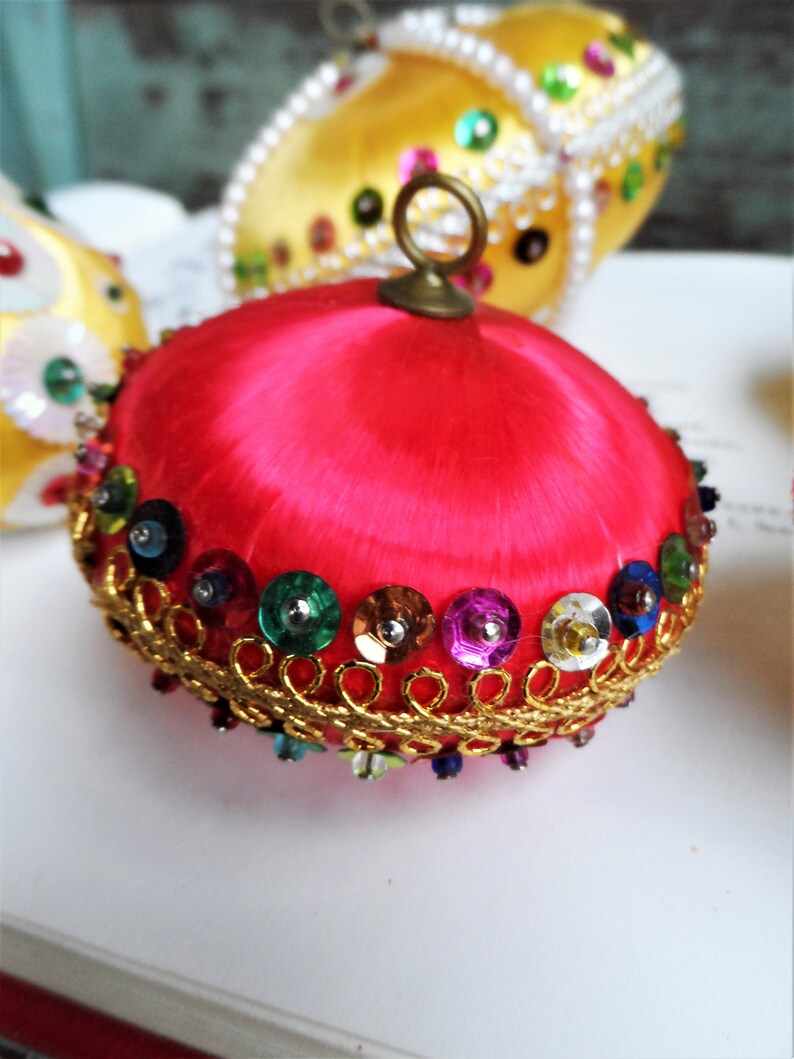 May include: A bright pink satin Christmas ornament with a gold trim and colorful sequins. The ornament has a loop at the top for hanging.