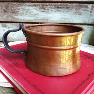 May include: A copper mug with a black handle sits on a red book. The mug has a cylindrical shape with a slightly flared rim and two horizontal bands. The handle is curved and attached to the side of the mug.