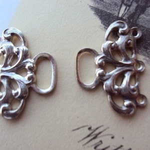 May include: Two silver-toned metal clasps with ornate, floral designs. Each clasp has a loop for attaching to a garment.