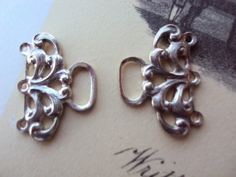 May include: Two silver-toned metal clasps with ornate, floral designs. Each clasp has a loop for attaching to a garment.