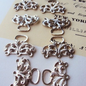 May include: Ten silver-toned metal decorative clasps with ornate floral designs. Each clasp has a loop on either side.
