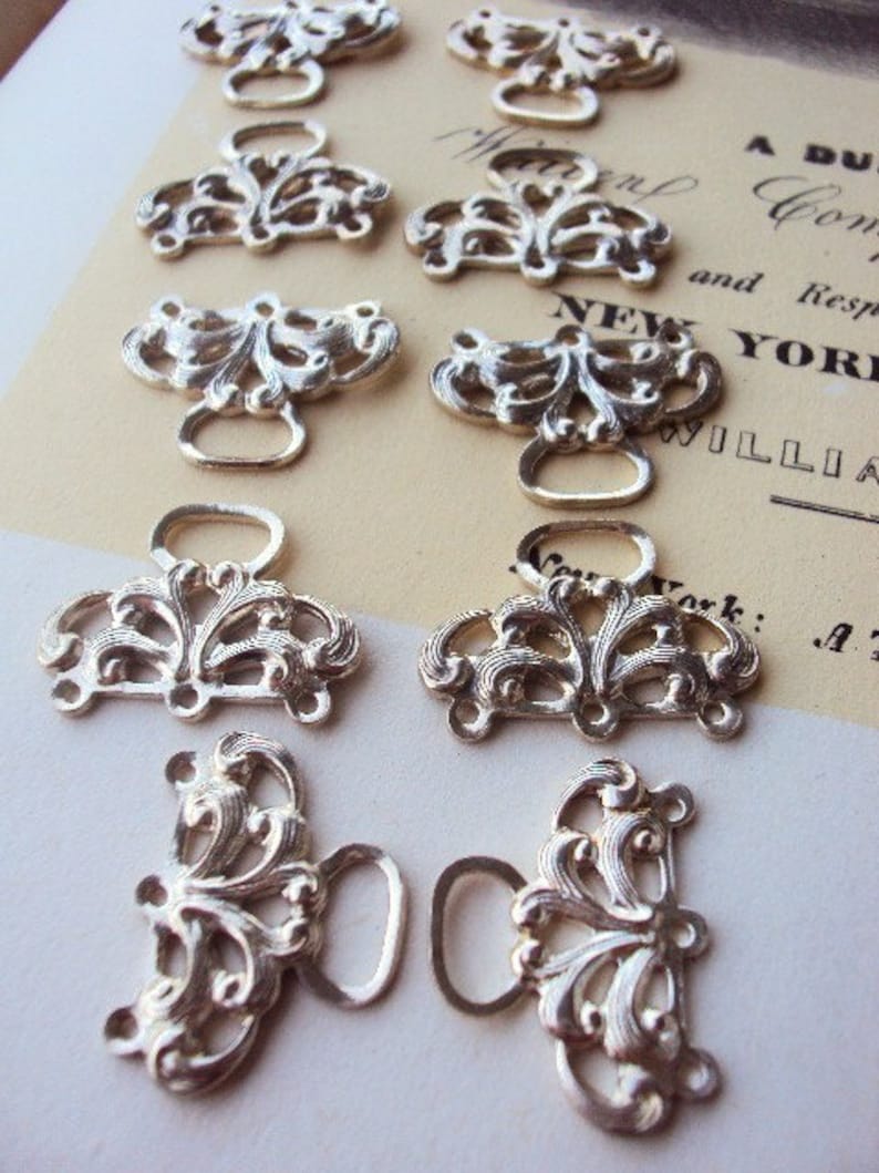 May include: Ten silver-toned metal decorative clasps with ornate floral designs. Each clasp has a loop on either side.