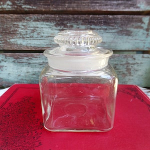 May include: A clear glass jar with a square body and a textured lid. The jar is sitting on a red surface.