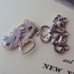 May include: Two silver metal decorative clasps with intricate designs. One clasp has a more ornate design with a floral motif, while the other has a more geometric design.