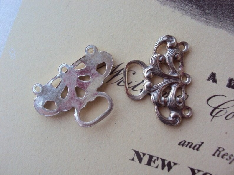 May include: Two silver metal decorative clasps with intricate designs. One clasp has a more ornate design with a floral motif, while the other has a more geometric design.