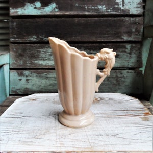May include: A pale pink ceramic pitcher with a fluted design and a sculpted handle in the shape of a lion's head.