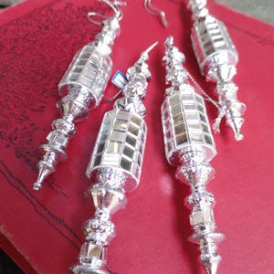 May include: Four silver Christmas ornaments with a mirrored, rectangular design and ornate detailing. Each ornament has a loop for hanging. The ornaments are displayed on a red surface.