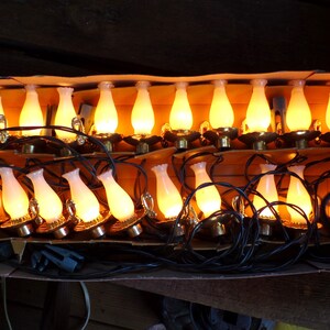 May include: A string of vintage lamp-shaped Christmas lights. Each light has a white glass shade and a brass-colored base, emitting a warm yellow glow. The lights are arranged in a cardboard box, with black wiring connecting them.
