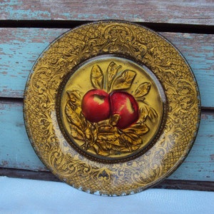 May include: A decorative plate with a gold filigree border and a central image of two red apples with green leaves. The plate has a vintage aesthetic and is set against a rustic wooden background.