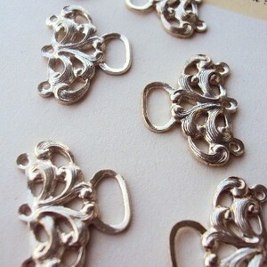 May include: Six silver-toned metal clasps with ornate designs. Each clasp has a loop on one side and a decorative, floral-inspired pattern on the other.