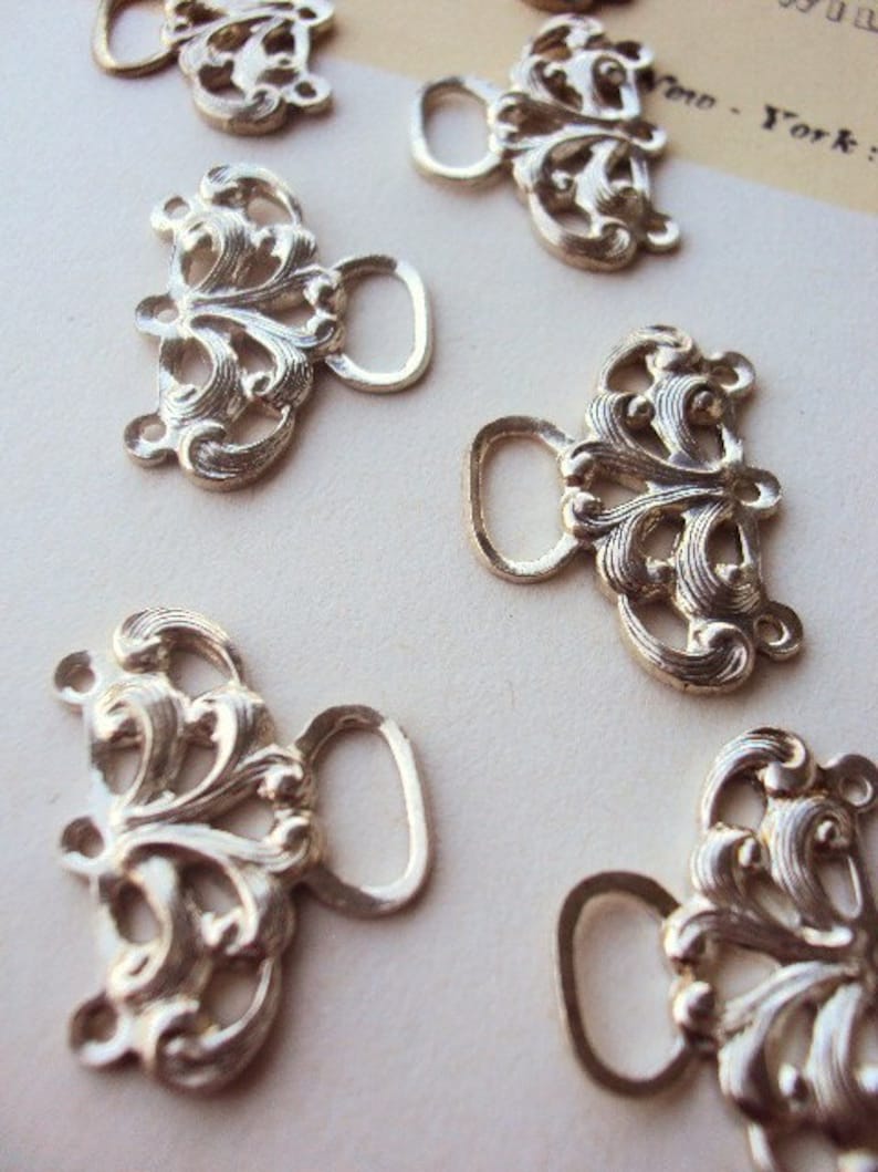 May include: Six silver-toned metal clasps with ornate designs. Each clasp has a loop on one side and a decorative, floral-inspired pattern on the other.