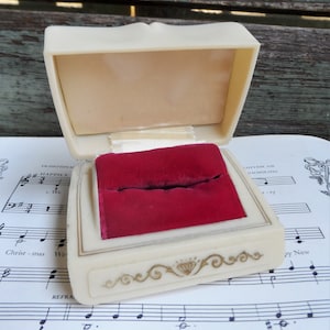May include: A vintage ring box made of off-white plastic with a red velvet interior. The box has a gold-colored design around the edge and a diamond-shaped design in the center.