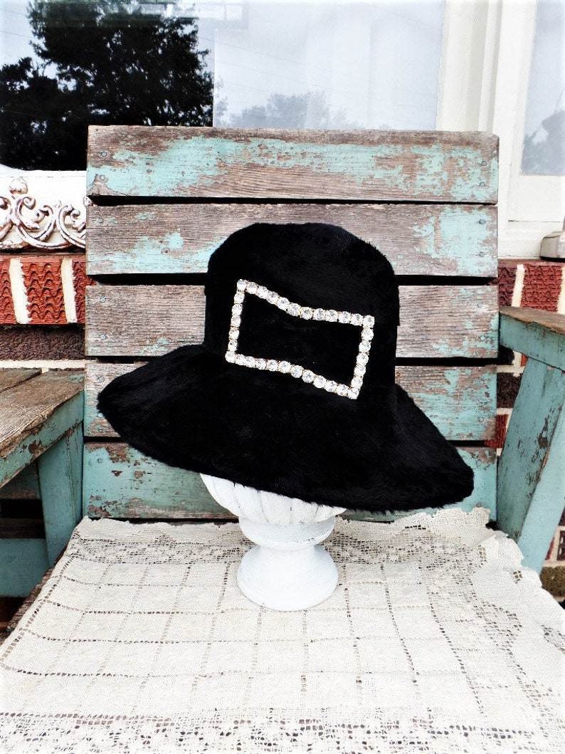 May include: A black wide-brimmed hat with a large, clear rhinestone embellishment in the shape of an envelope.