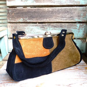May include: A color-blocked handbag crafted from suede panels in black, burnt orange, and olive green. The bag features a gold-tone metal frame with a kiss-lock closure and a black suede handle attached with gold-tone rivets on each side.