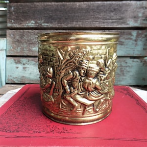 May include: A gold metal planter with an embossed design of people working in a medieval village. The planter is sitting on a red book.