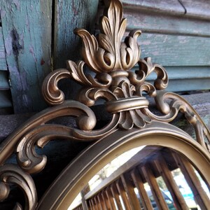 May include: A gold-toned oval mirror with an ornate, decorative frame. The frame features a large, intricate floral design with a central crest.