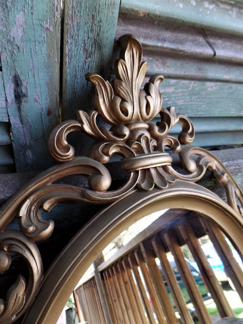May include: A gold-toned oval mirror with an ornate, decorative frame. The frame features a large, intricate floral design with a central crest.