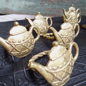 Könnte beinhalten: Nahaufnahme von mehreren goldfarbenen Serviettenringen in Teekannenform. Jeder Ring hat ein detailliertes Design mit Griff, Ausguss und Deckel. Die Serviettenringe sind auf einer strukturierten schwarzen Oberfläche angeordnet.