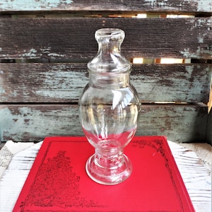May include: A clear glass jar with a lid, sitting on a red book with a black and white illustration. The jar has a round body and a wide base.