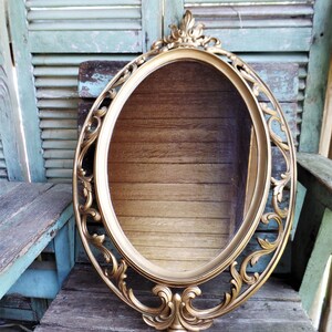 May include: A gold oval mirror with an ornate, carved frame. The frame has a decorative design with scrolls and floral motifs. The mirror is hanging on a wooden wall.