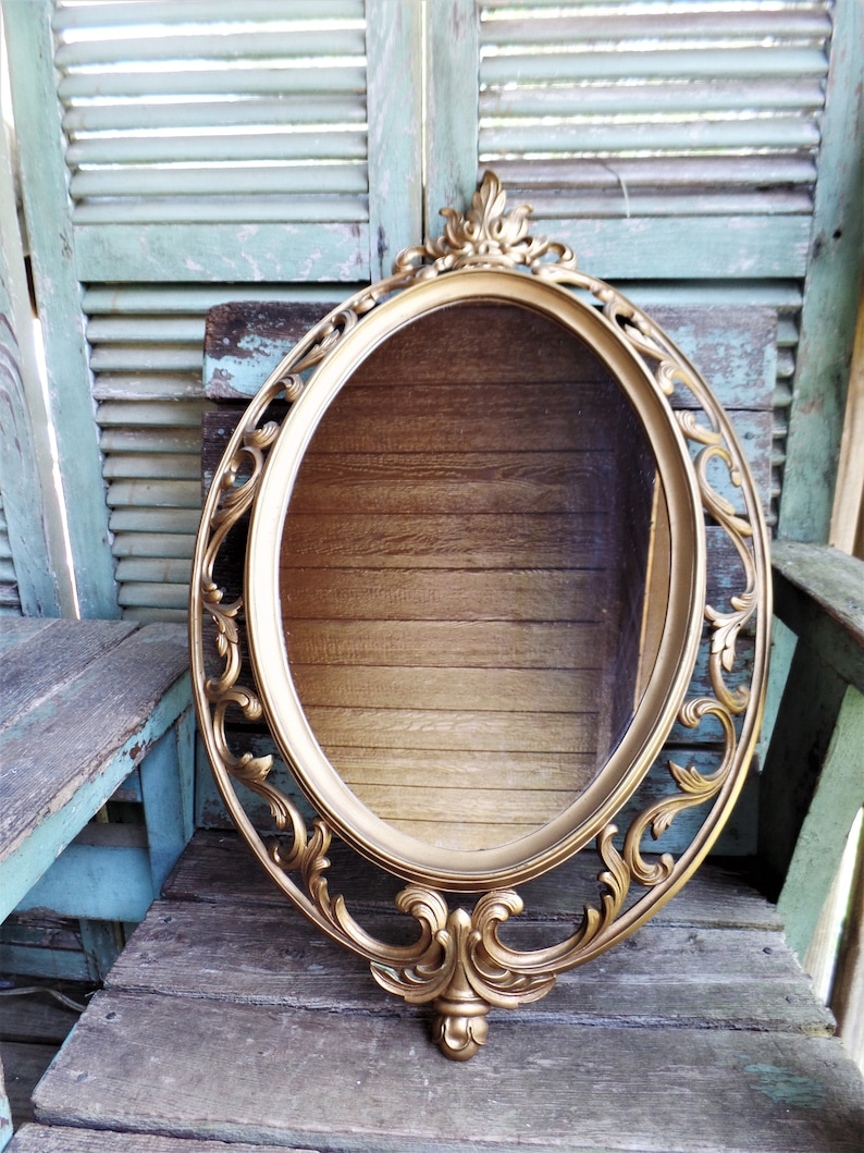 May include: A gold oval mirror with an ornate, carved frame. The frame has a decorative design with scrolls and floral motifs. The mirror is hanging on a wooden wall.