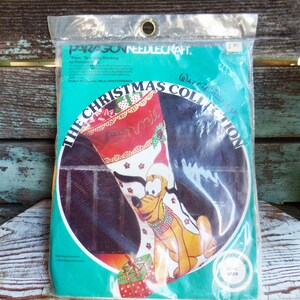 May include: A plastic package containing a needlepoint kit featuring a Christmas stocking with a cartoon image of Pluto the dog. The stocking is red and green with white polka dots and the text "The Christmas Collection" and "What's New,  Pluto?" The kit is from Paragon Needlecraft.