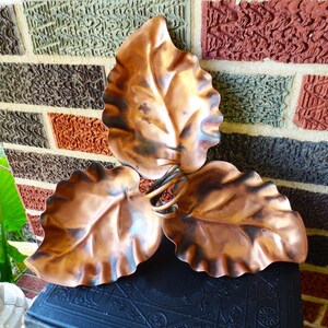 May include: A decorative metal dish featuring three interconnected copper-colored leaves with ruffled edges and dark, shaded accents. The leaves are joined at the center by a coiled metal stem, creating a tri-lobed, organic shape.
