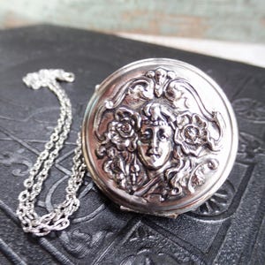May include: Silver-toned round pendant with a detailed relief of a woman's face surrounded by flowers and foliage. The pendant is attached to a delicate chain. The piece is likely vintage or antique.