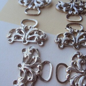 May include: Eight silver-toned metal clasps with ornate floral designs and a loop for fastening. The clasps are arranged in two rows on a white surface.