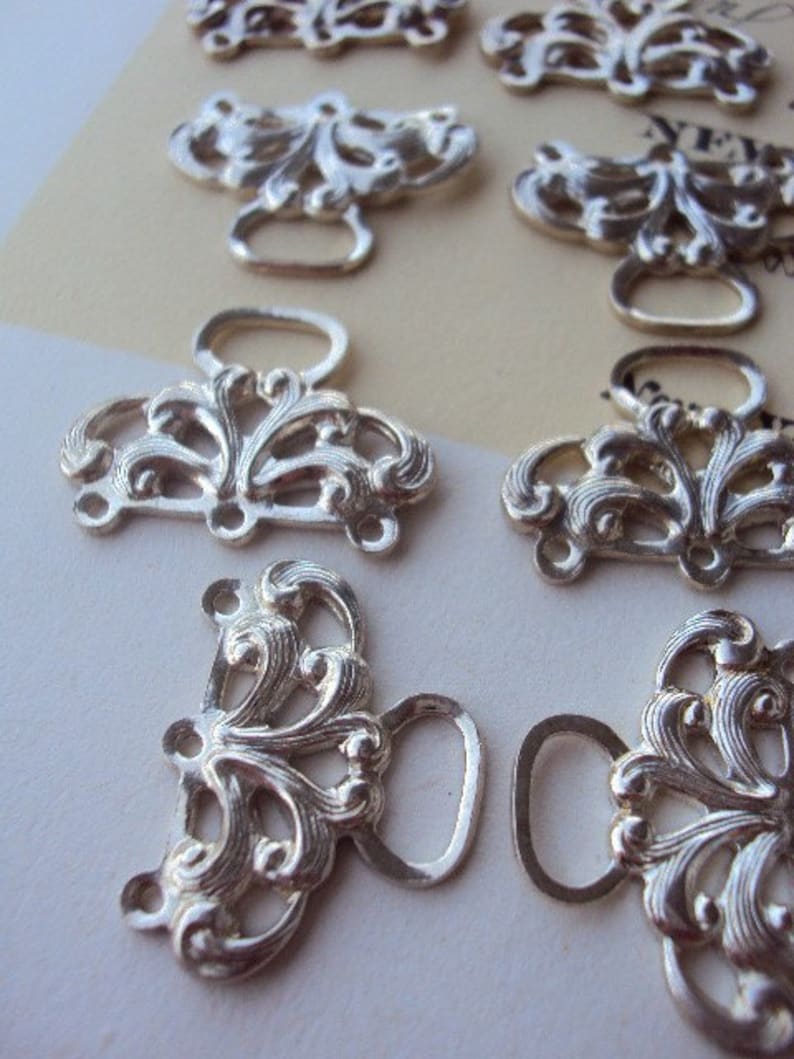 May include: Eight silver-toned metal clasps with ornate floral designs and a loop for fastening. The clasps are arranged in two rows on a white surface.