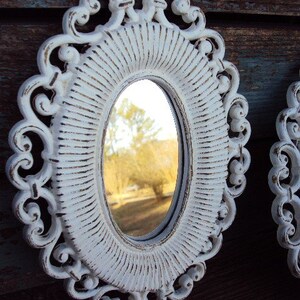 May include: White oval mirror with a decorative, ornate frame. The frame has a distressed, aged look with a white paint finish. The mirror reflects a view of trees and a grassy area.