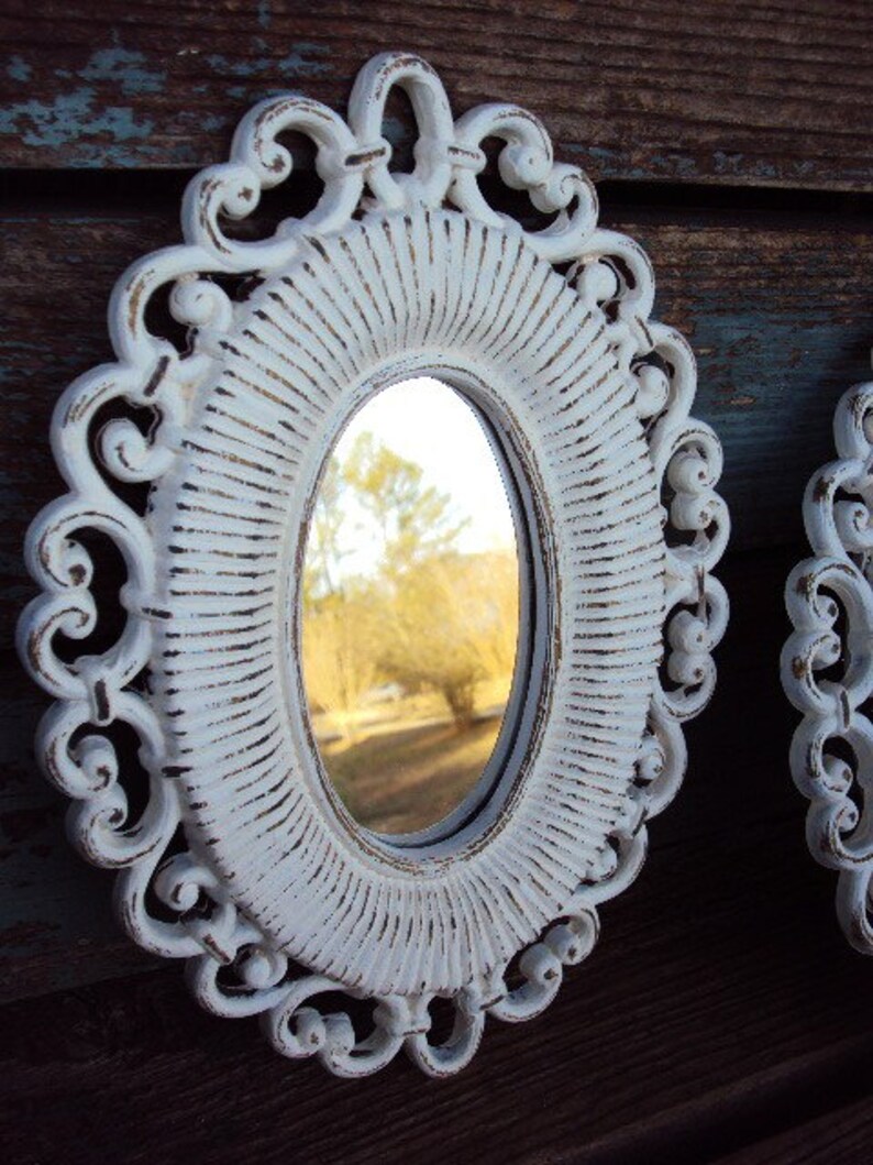 May include: White oval mirror with a decorative, ornate frame. The frame has a distressed, aged look with a white paint finish. The mirror reflects a view of trees and a grassy area.