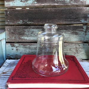 May include: A clear glass jar with a wide mouth and a bell-shaped body sits on top of a red book with black lettering. The jar is empty and has a smooth, rounded surface.
